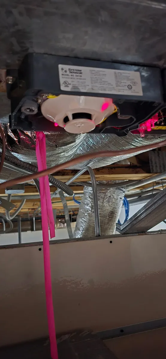 A System Sensor D4120 duct smoke detector mounted on the underside of a duct in a ceiling space, red alarm LED lit