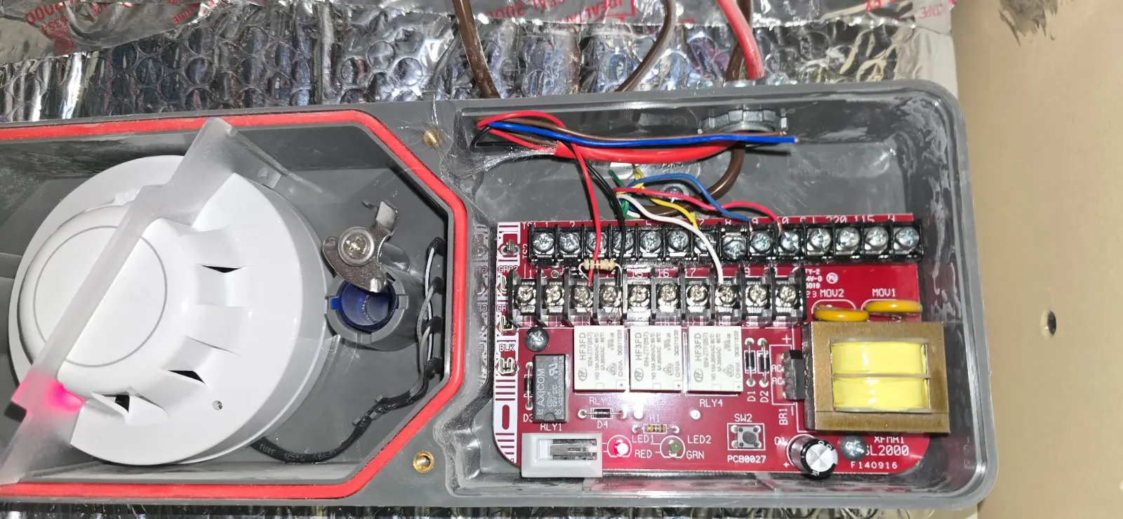 An SL-2000 duct smoke detector housing opened, showing the sensing head at left and the relay board and terminal strip at right